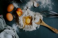 Top view pile of flour on table with broken egg arranged with wooden spoon and metal corolla during preparation of homemade dough