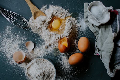 From above of broken eggs on flour pile scattered on table near salt sack and kitchenware