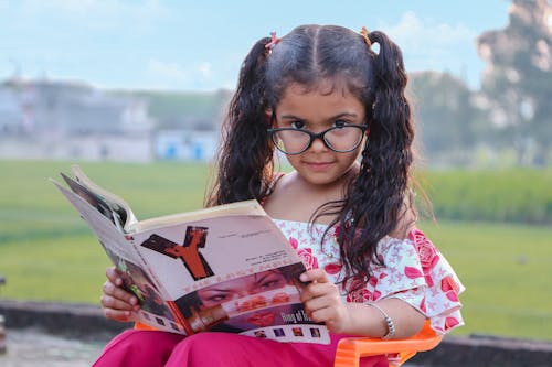 Free Funny ethnic little girl with curly pigtails and eyeglasses sitting on chair in park and reading magazine with interest Stock Photo