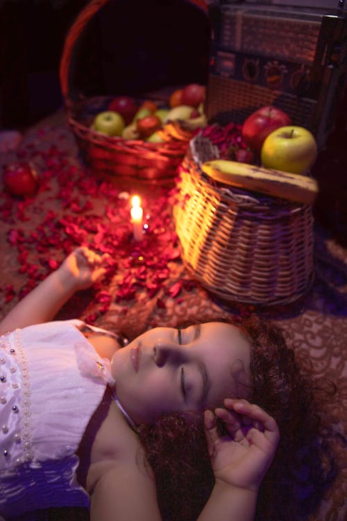 Free From above of cute little girl with curly hair lying on floor and sleeping near burning candle and baskets with assorted fresh fruits Stock Photo