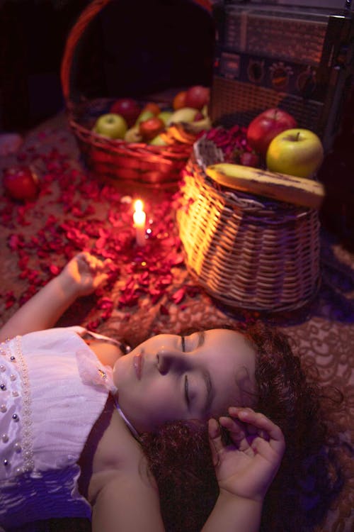 From above of cute little girl with curly hair lying on floor and sleeping near burning candle and baskets with assorted fresh fruits