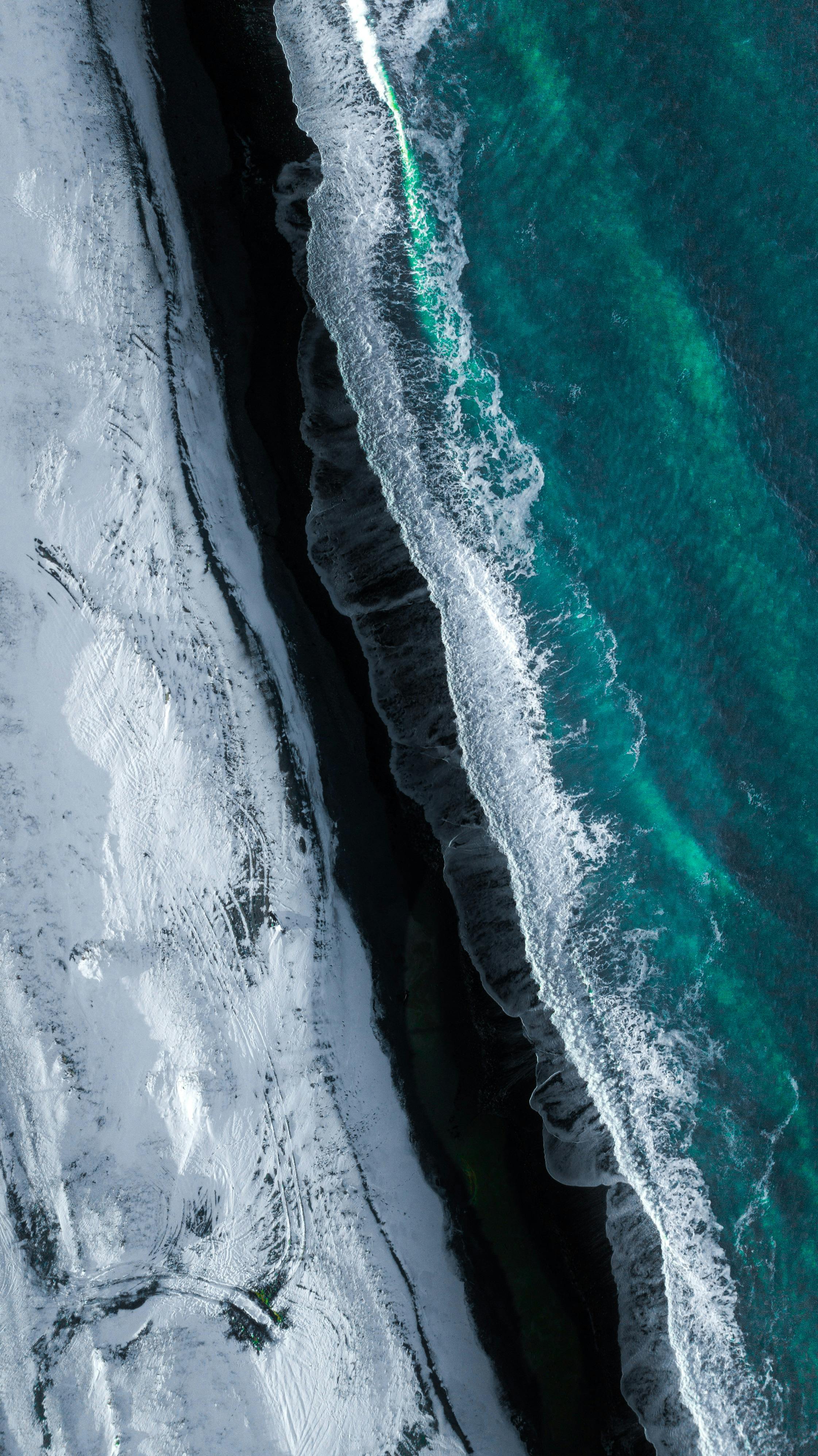 Picturesque aerial view of foamy waves of turquoise ocean washing black sandy beach covered with snow