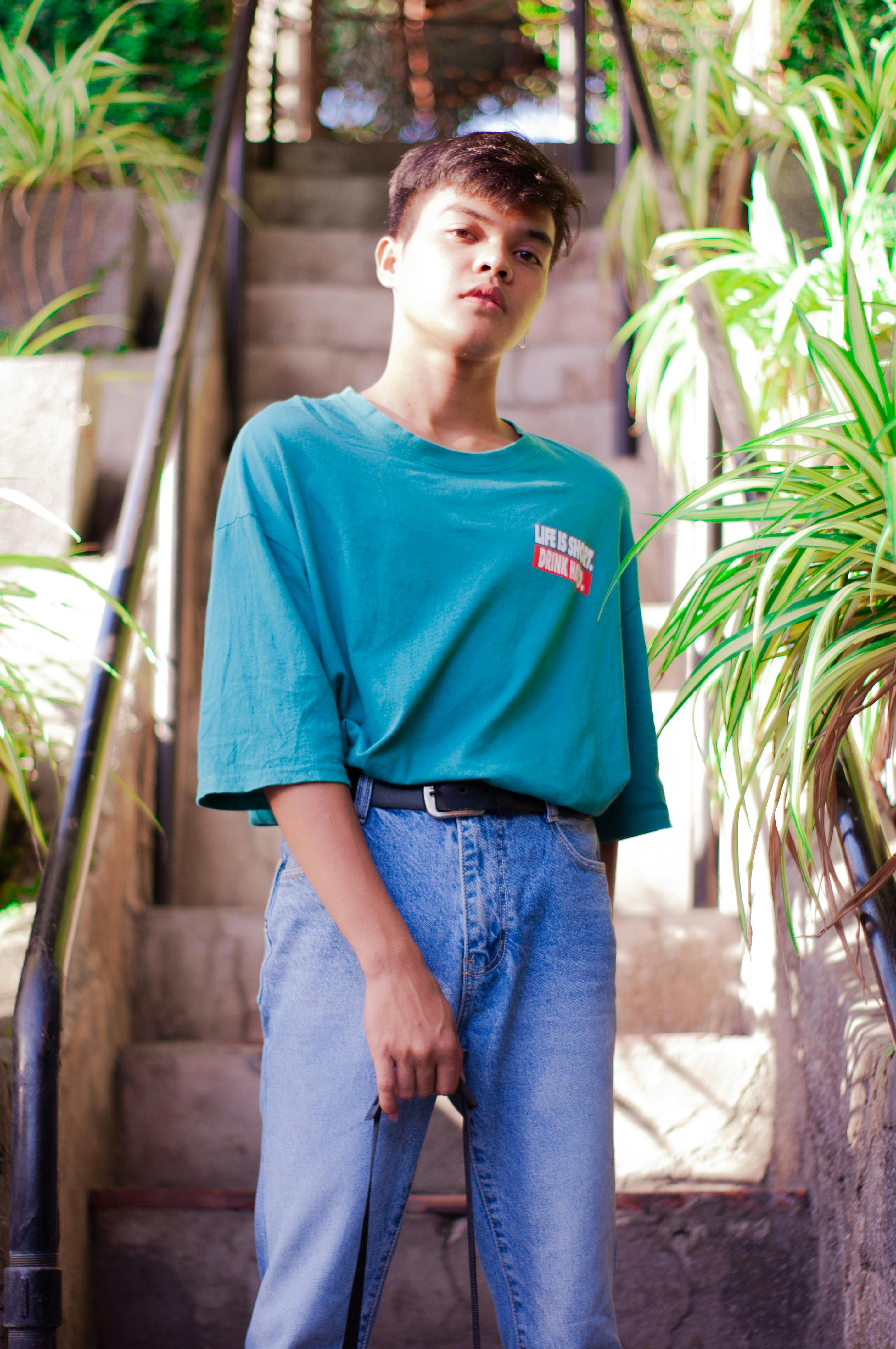 Low angle of confident ethnic young male millennial in stylish outfit standing near staircase decorated with green plants and looking at camera