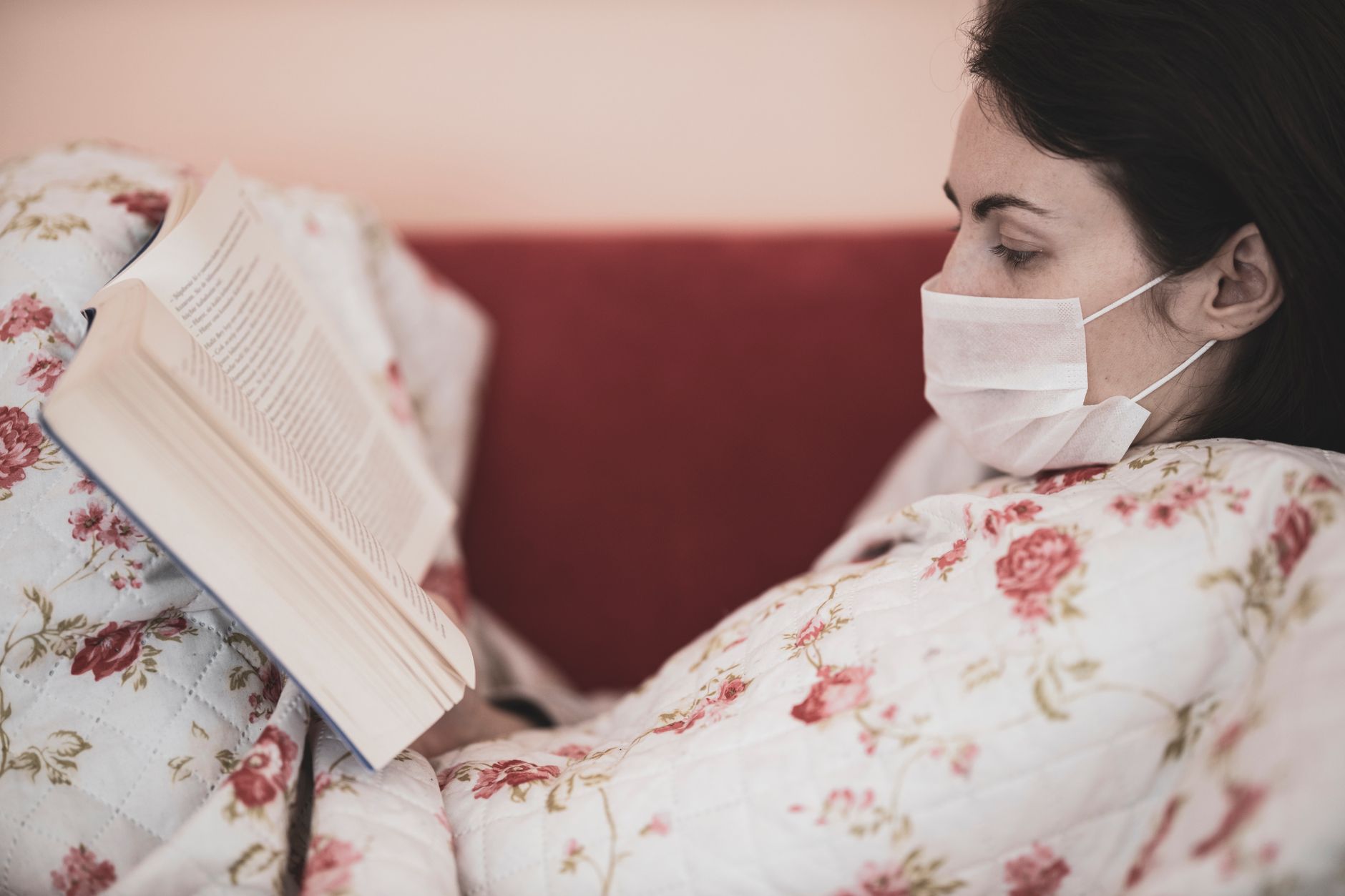 long covid recovery — A woman wearing a facemask reads a book while wrapped in a floral blanket indoors.