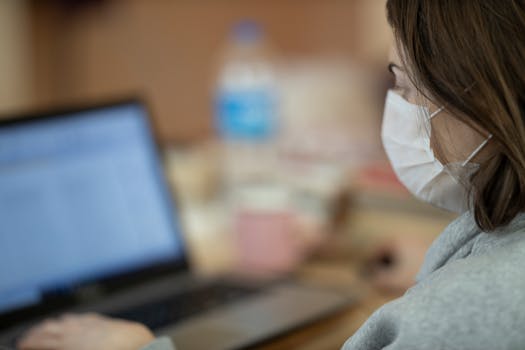 Close-up of a person working remotely on a laptop, wearing a facemask for safety.