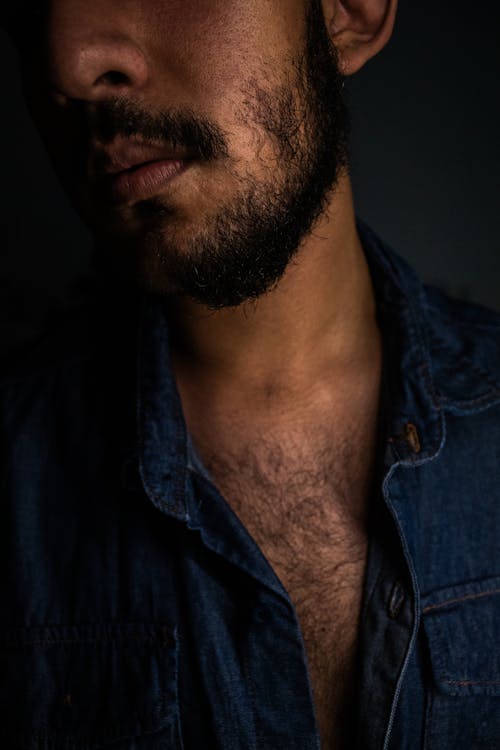 Faceless young ethnic male in denim shirt with beard and hairy chest on dark background