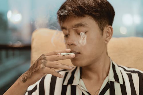Thoughtful young ethnic male in striped shirt with tattoo on hand smoking while resting in chair in street cafe on blurred background