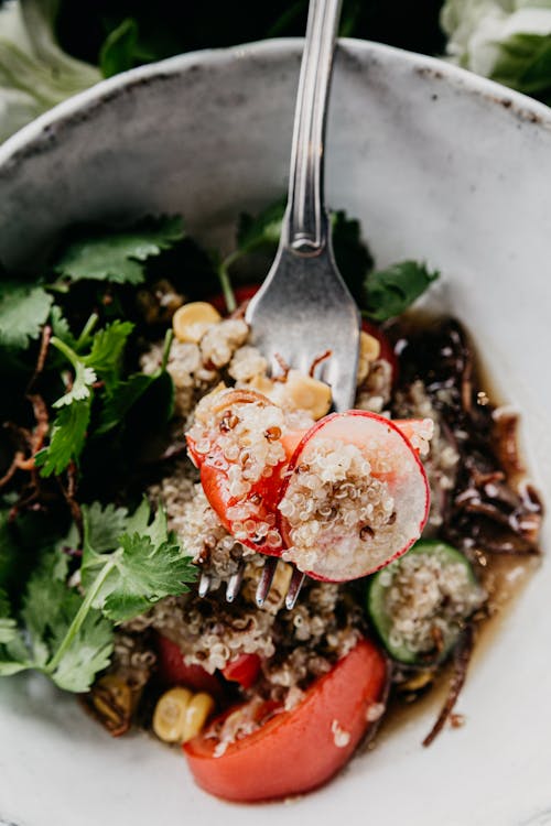 Top view of delicious healthy couscous salad with vegetables and herbs with balsamic dressing placed in white ceramic bowl with stainless steel fork