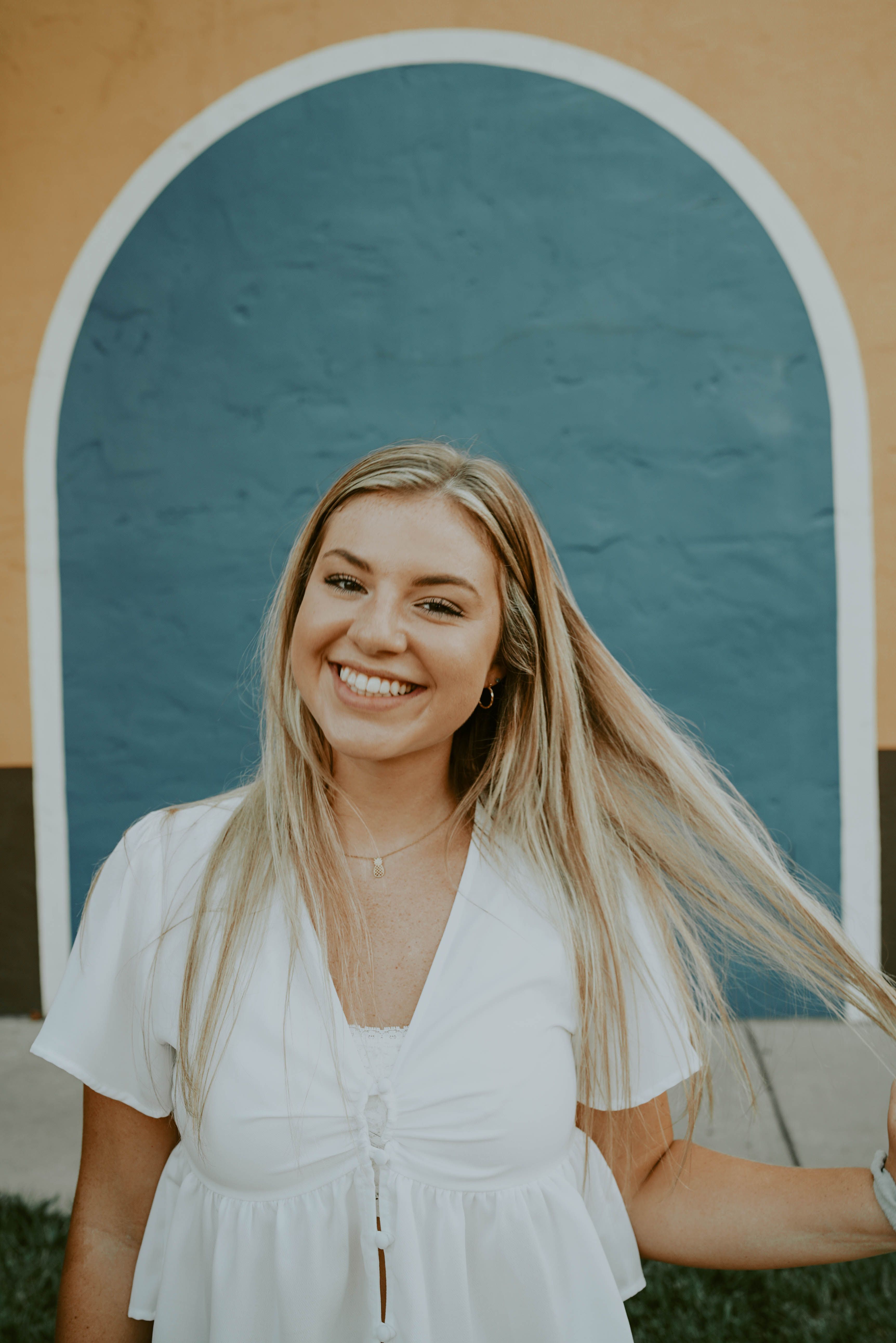 Young cheerful female touching hair while smiling brightly and standing against bright building