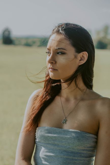 Dreamy young female with bare shoulders and flowing hair thoughtfully looking away while standing in meadow