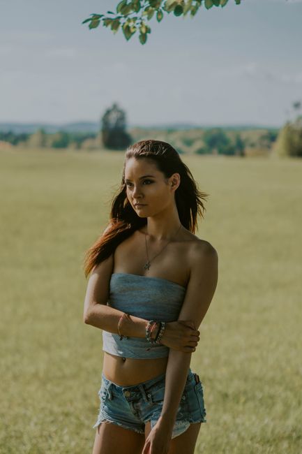 Calm young female in summer outfit standing in field in countryside and thinking while looking away