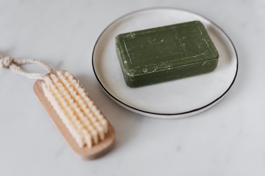 Close-up of a green soap bar on a dish with a wooden brush on a marble surface.