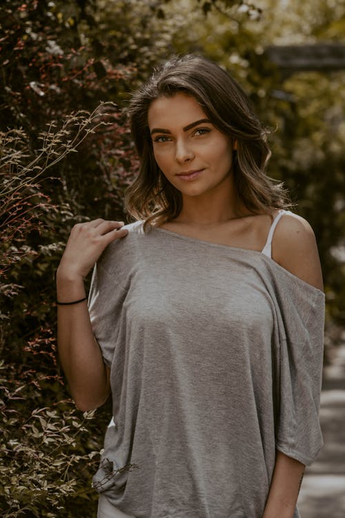 Young female standing against greenery and gently touching shoulder while looking at camera