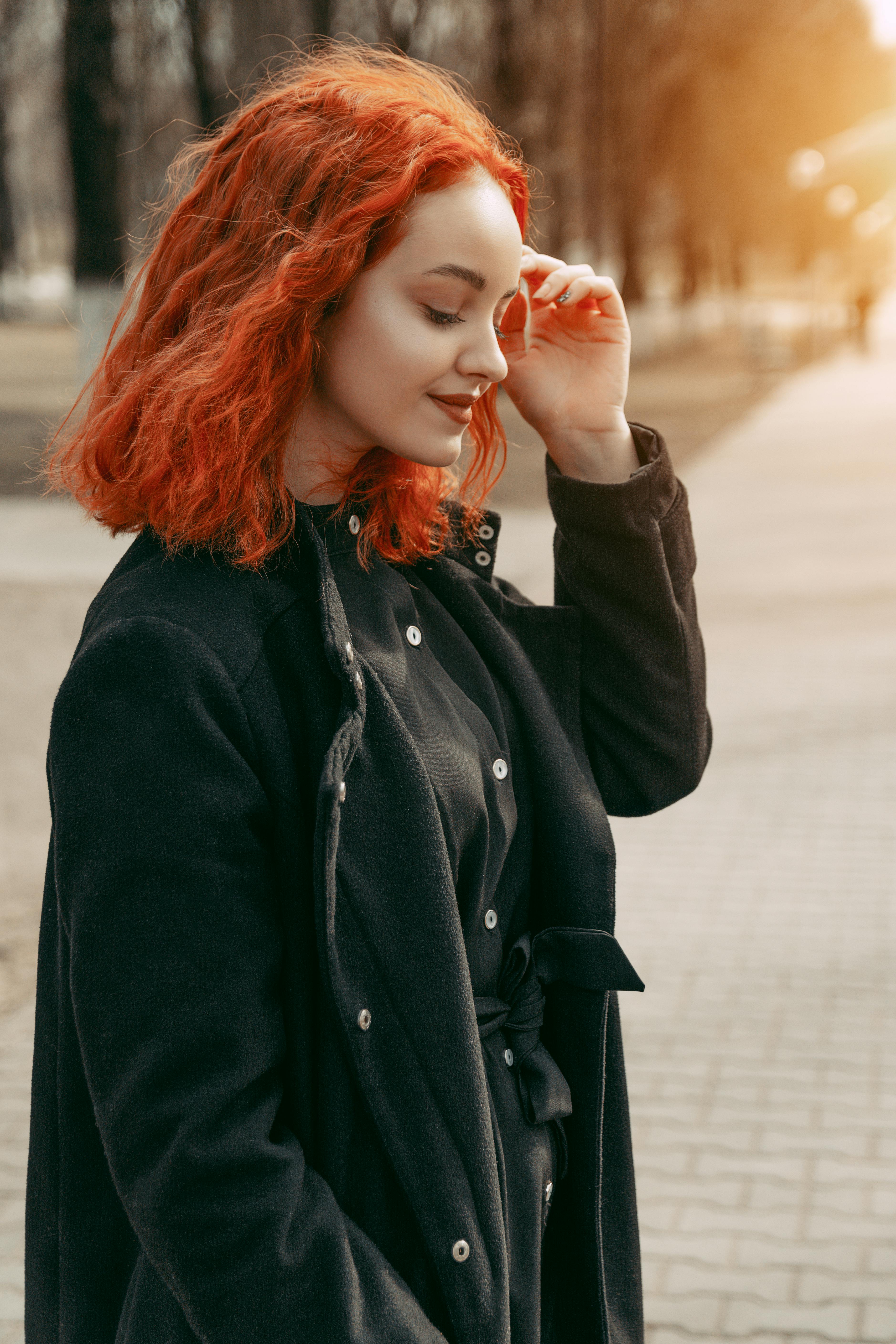 A young woman with vibrant red hair smiling while outdoors wearing a black coat.