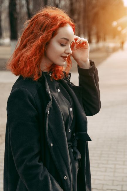 A young woman with vibrant red hair smiling while outdoors wearing a black coat.