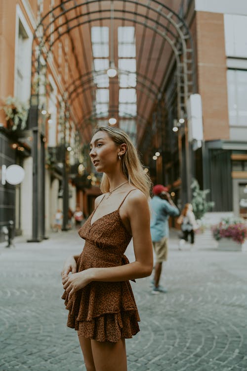 Side view of young thoughtful female in summer dress standing against modern building with arched ceiling