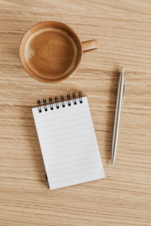 Top view of beige ceramic cup of aromatic espresso placed near ring bound notebook with blank sheet and silver pen on wooden tabletop