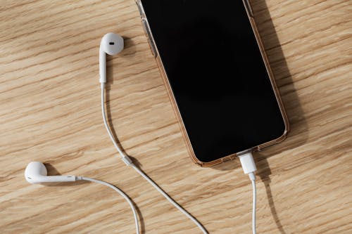 From above composition of white wired earphones and modern mobile phone with blank screen and connected charging cable placed on wooden surface in daylight
