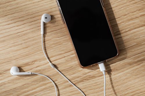 Free From above composition of white wired earphones and modern mobile phone with blank screen and connected charging cable placed on wooden surface in daylight Stock Photo