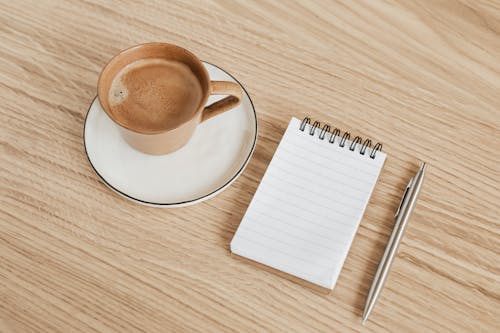 Free From above of ceramic cup of hot espresso on white saucer placed near opened ring bound notebook and stylish silver pen on wooden surface Stock Photo