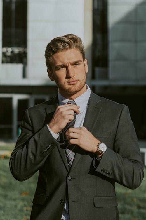Serious confident male entrepreneur wearing classy suit and wristwatch holding tie while thoughtfully looking away