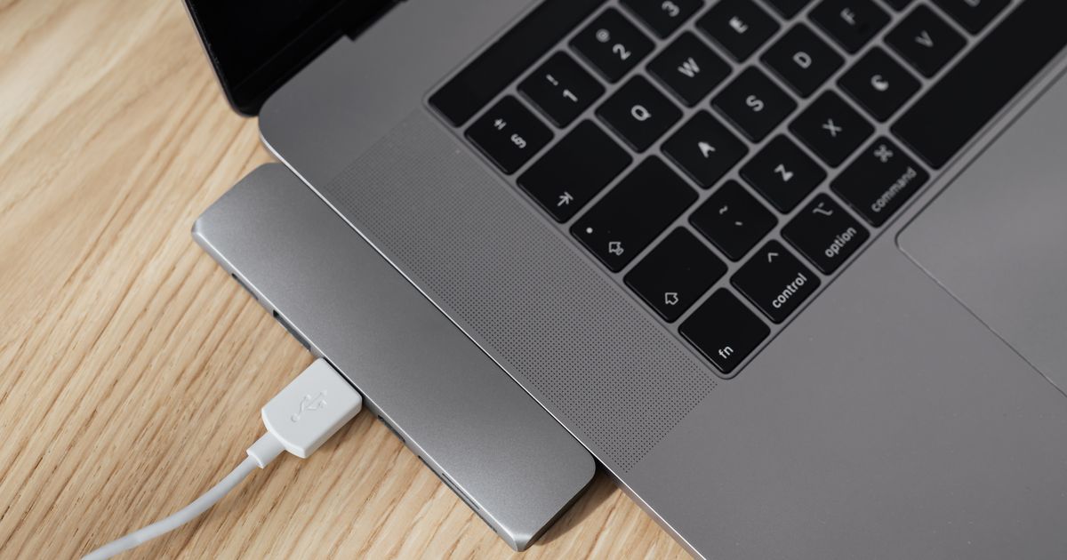A slim space-grey USB-C hub plugged into the side of a laptop on a light oak desk, a white USB cable in one of its ports