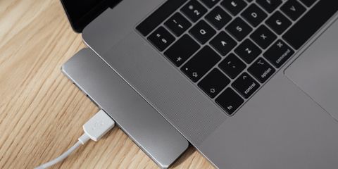 A slim space-gray USB-C hub plugged into the side of a laptop on a light oak desk, a white USB cable in one of its ports