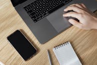 木制桌子上，笔记本电脑触控板的手部鸟瞰图旁，放着智能手机和记事本