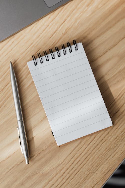 From above of small opened notepad and stylish silver pen placed on wooden table near laptop