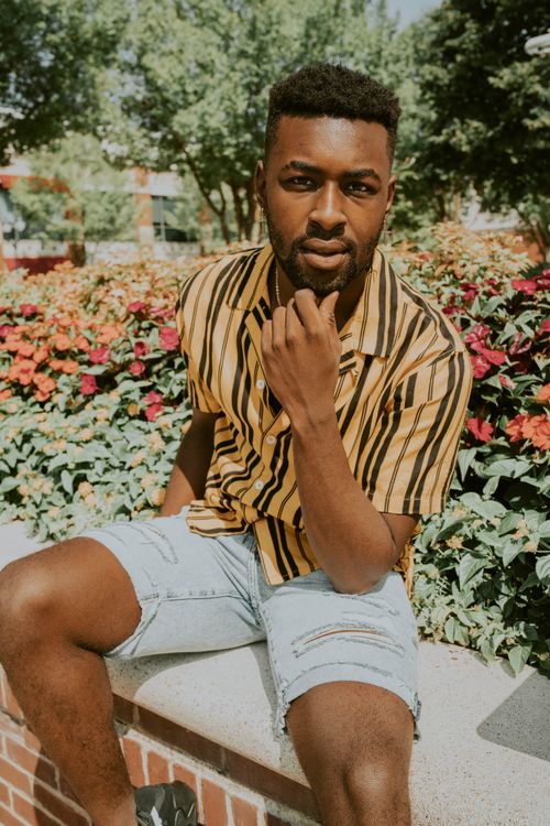 Free Young stylish black male in modern wear touching unshaven chin while sitting behind blooming flowers and looking at camera Stock Photo