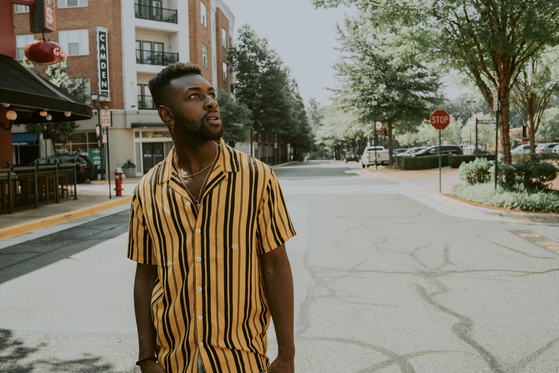 Young stylish dreamy African American guy in bright striped shirt and creative haircut looking up on asphalt roadway