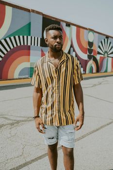 Young wistful African American male in bright ornamental shirt standing on road behind graffiti wall and looking away