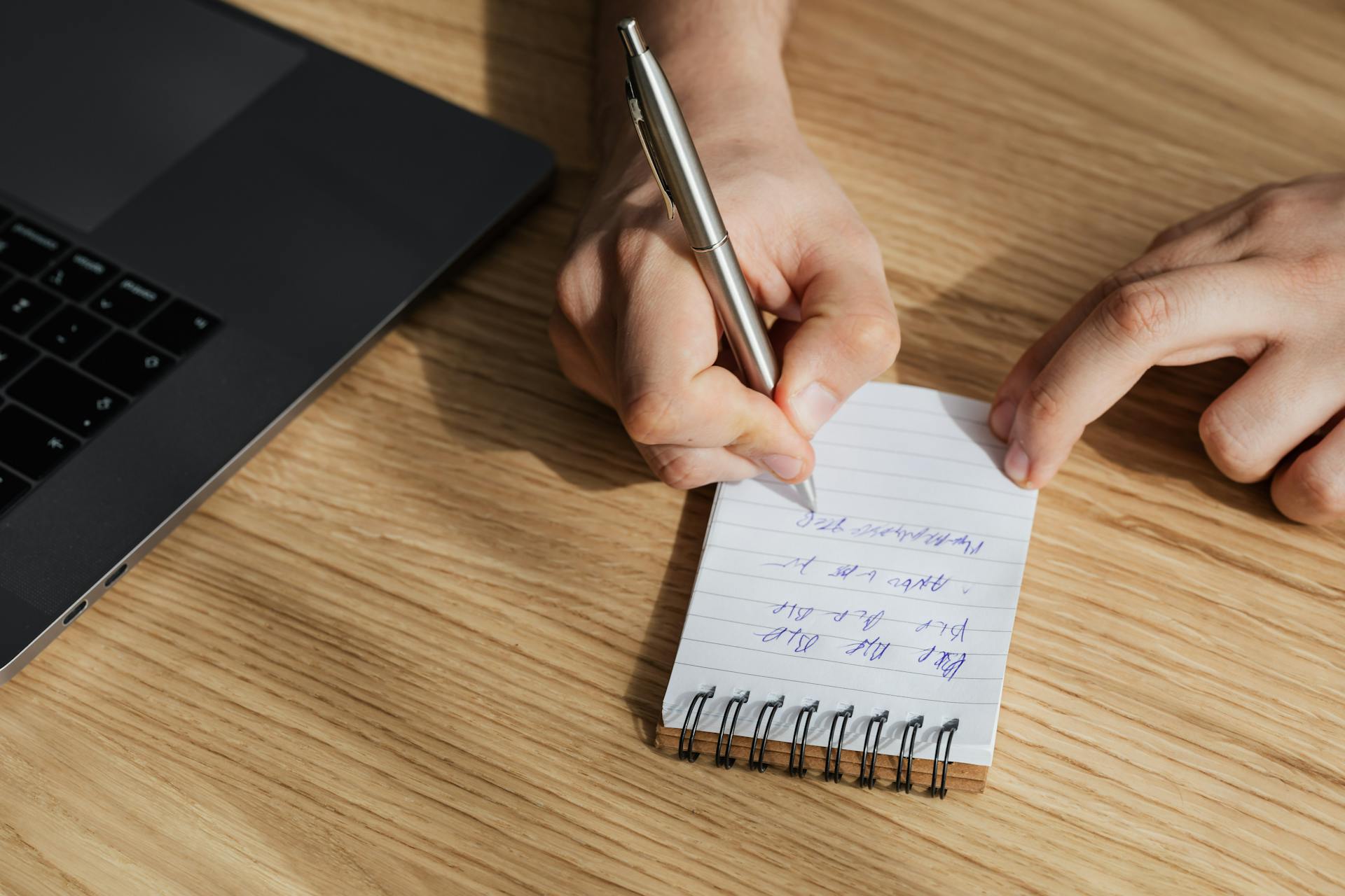 From above of crop unrecognizable man writing out information in spiral notebook while working on business plan on laptop