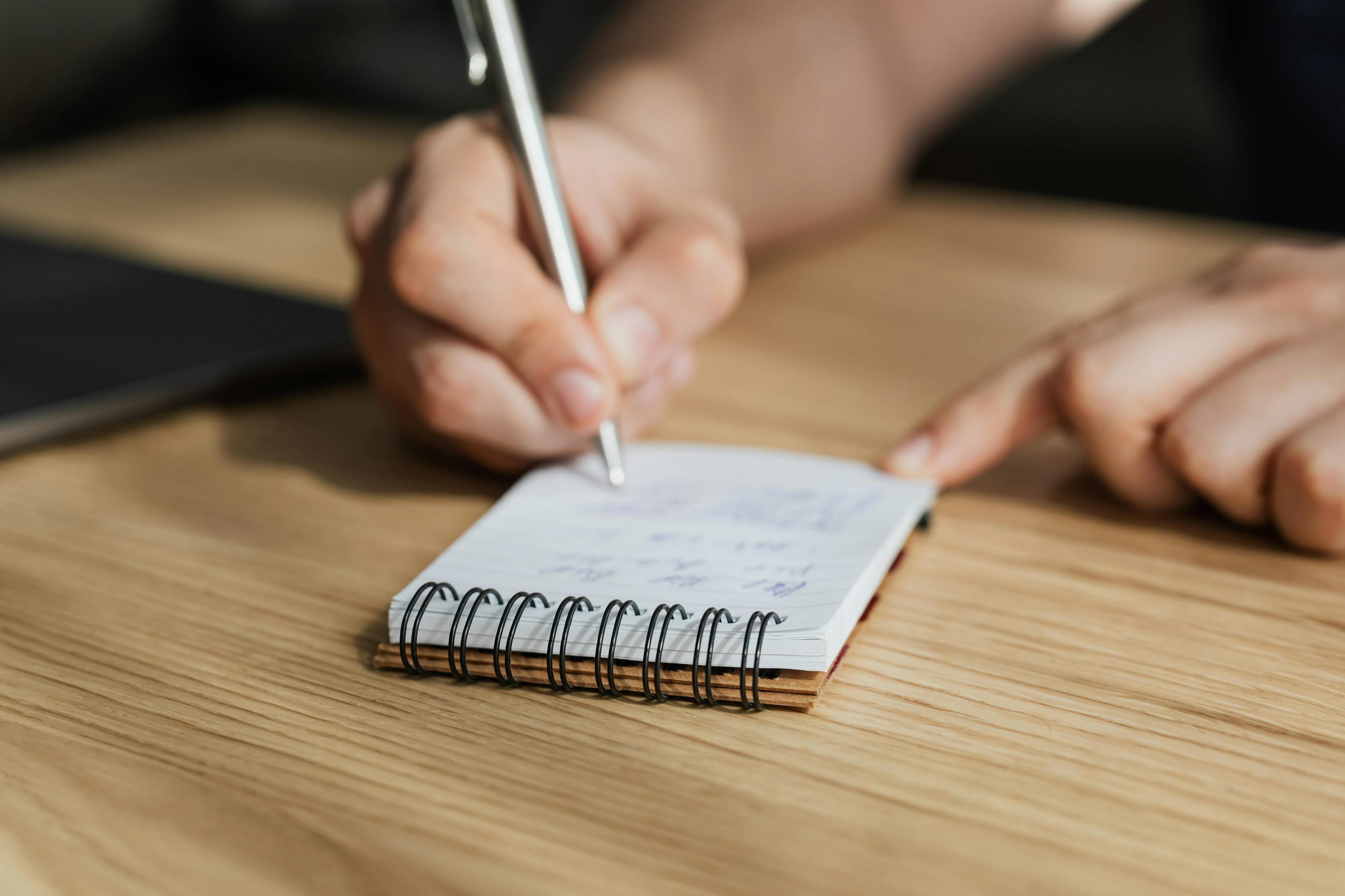  A person is seen taking notes in a spiral notebook while sitting at a wooden table.