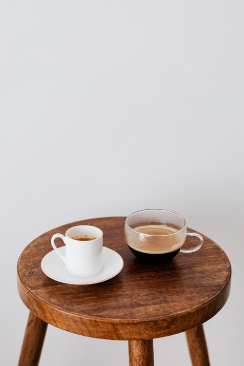 Free Fresh coffee in ceramic white cup and wide glass cup placed on round wooden table near white wall Stock Photo
