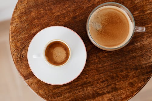From above of coffee in white cup served on saucer and of coffee in wide glass cup placed on round wooden table