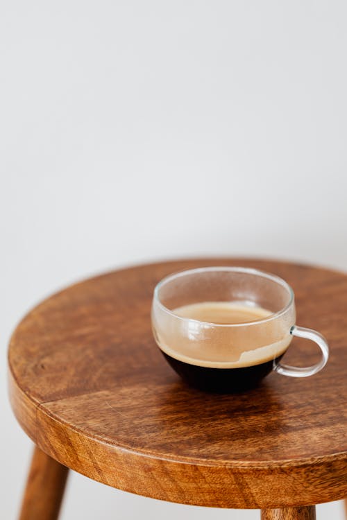 Black coffee in wide glass cup placed on round wooden table near white wall
