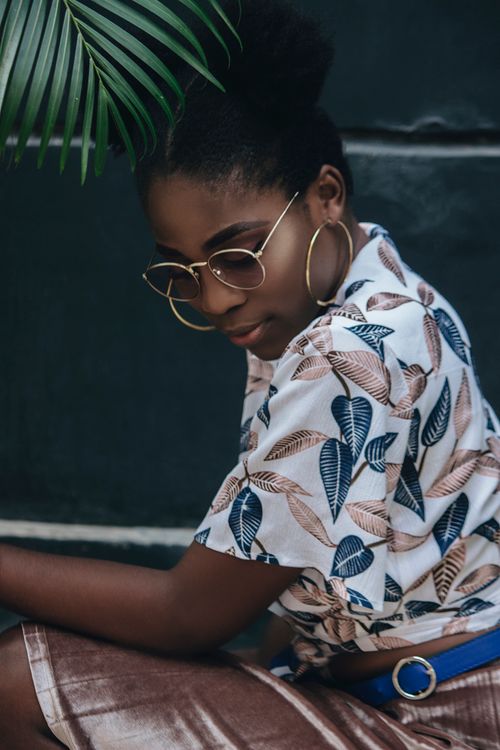 Free Portrait of a stylish woman wearing sunglasses and a patterned shirt, sitting outdoors amongst greenery. Stock Photo