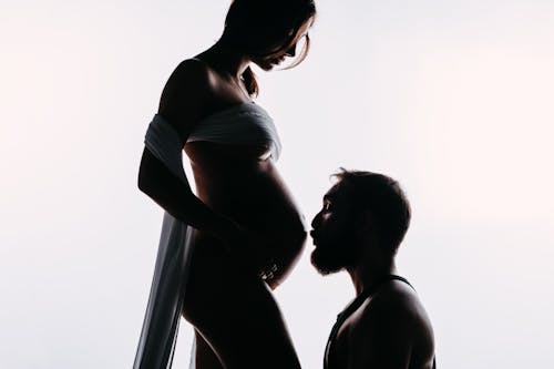 Free Side view of bearded male before kissing belly of crop anonymous expectant female beloved on white background Stock Photo