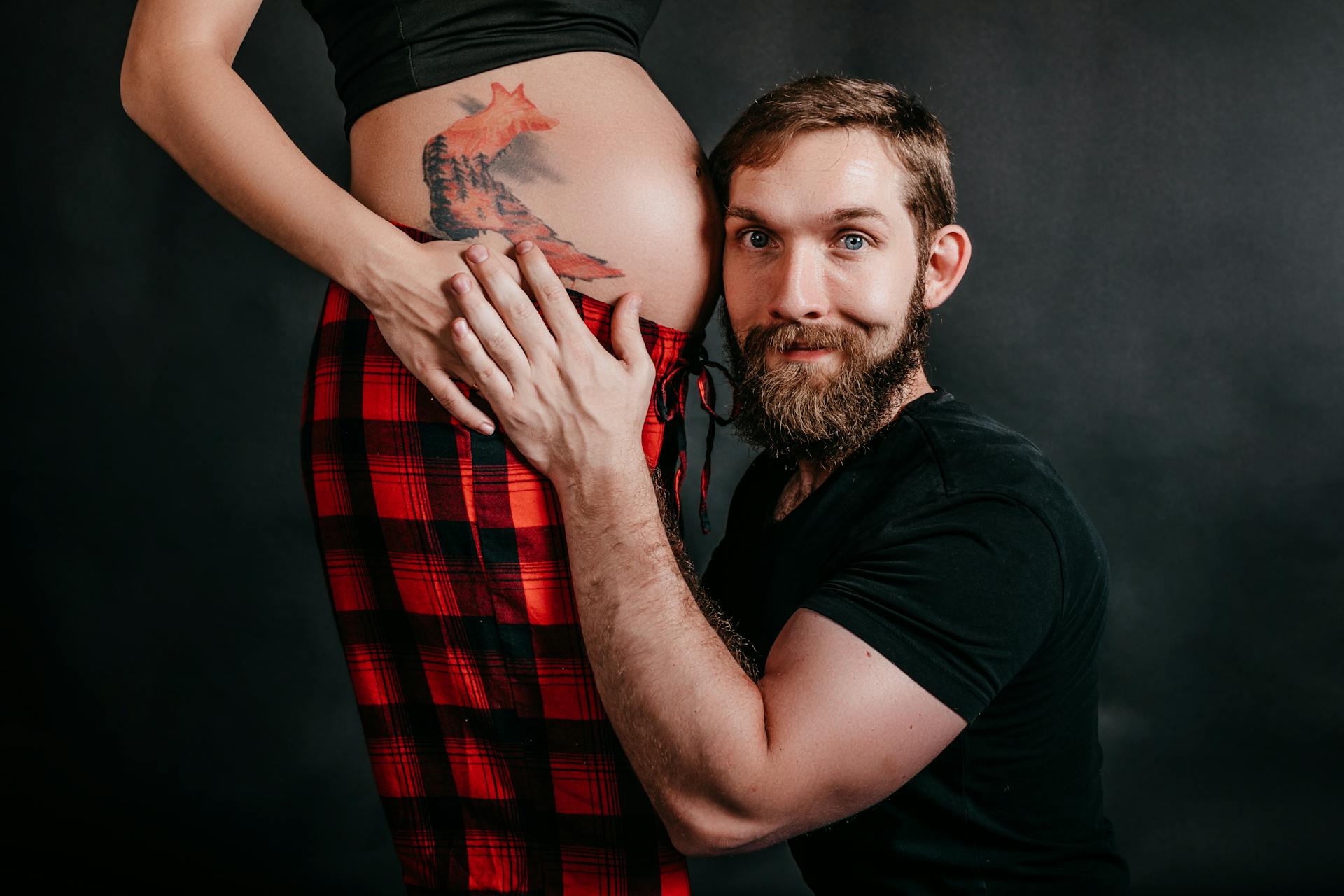 Side view of smiling unshaven male embracing belly of crop anonymous tattooed expectant girlfriend while looking at camera