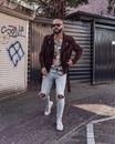 一位戴着墨镜、身穿麂皮夹克和破洞牛仔裤的时尚秃头男子，走在涂鸦遍布的城市街道上