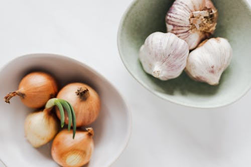 Top view of ceramic bowls of garlic bulbs and onions with green sprouts on white marble table