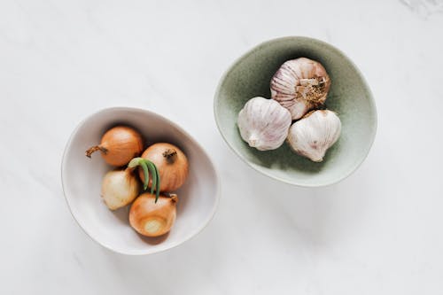 Top view of unpeeled raw yellow onion in white bowl and unpeeled raw aromatic garlic in light green bowl placed on marble table