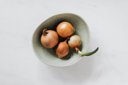 Top view of light green bowl with unpeeled raw organic yellow onion bulbs and one regrowing onion placed on white marble tabletop