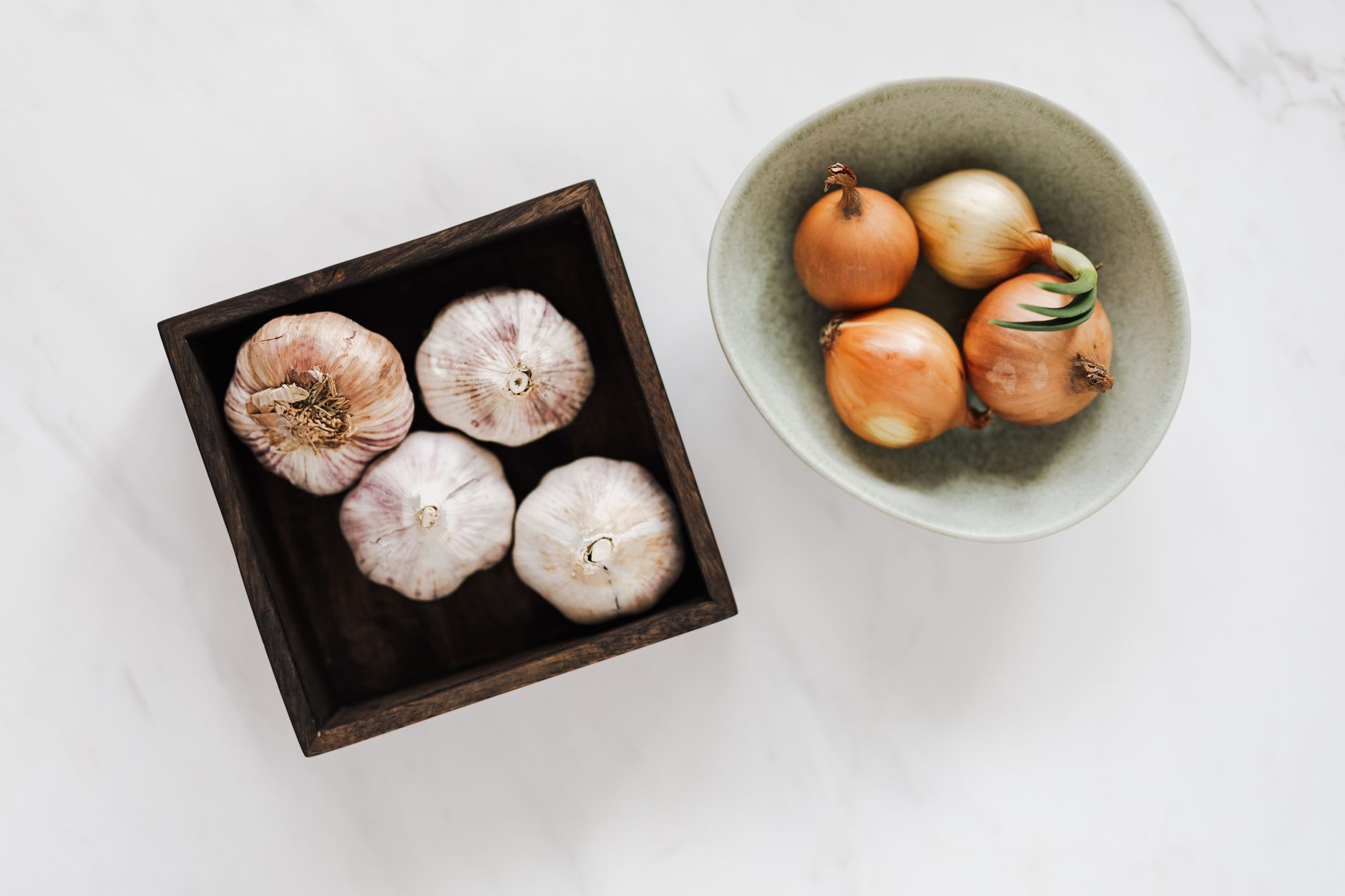 Overhead of square pot with fresh unpeeled garlic near light green bowl with raw yellow onions against white marble surface