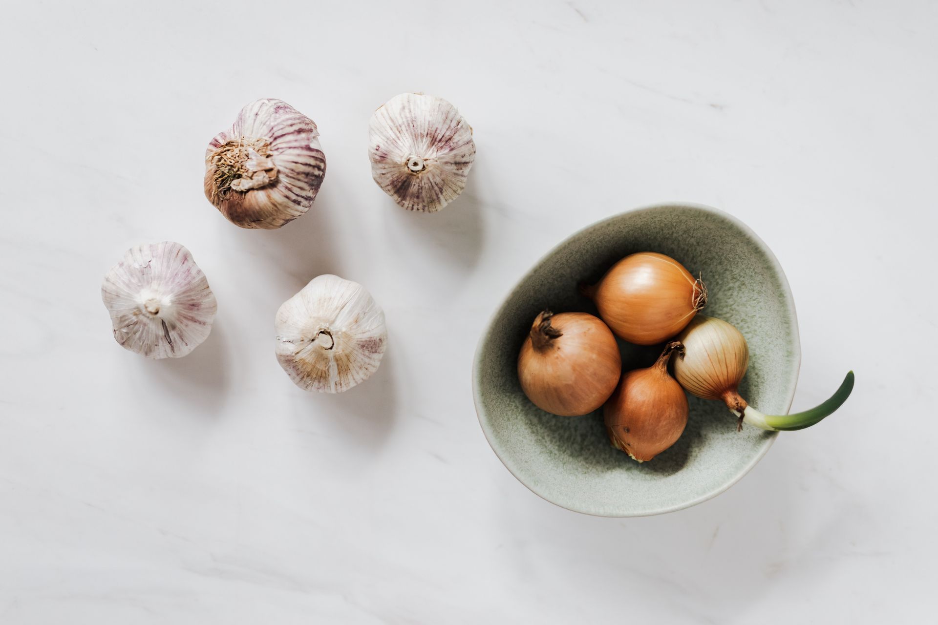 Top view of ceramic bowl filled with raw unpeeled yellow onions and one regrowing bulb placed near aromatic garlic laying on white marble tabletop