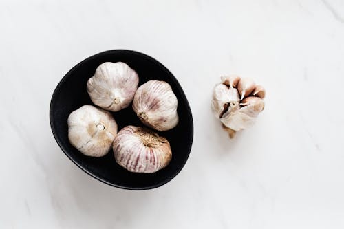 Top view of aromatic raw unpeeled garlic in black bowl near half peeled garlic bulb placed on white marble table