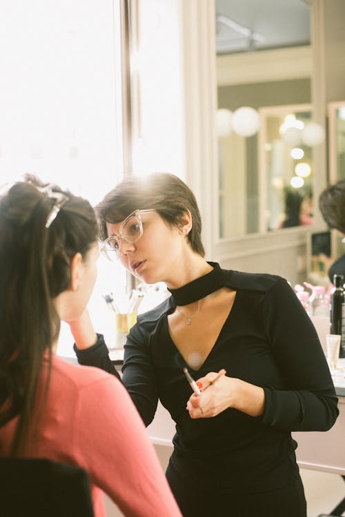Focused visagiste in black casual outfit and eyeglasses applying makeup on female customer while standing against cozy dressing table with big mirror and lamps
