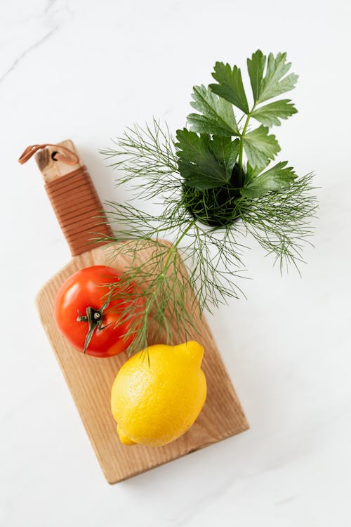 Overhead view of raw tomato and lemon lying on cutting board with dill and parsley in cup beside on marble table