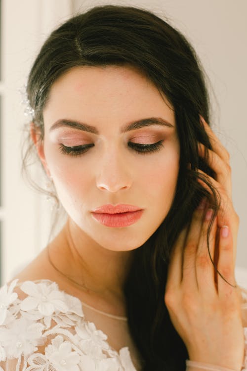 Young dark haired bride with trendy makeup in white dress touching hair and looking down while posing in studio